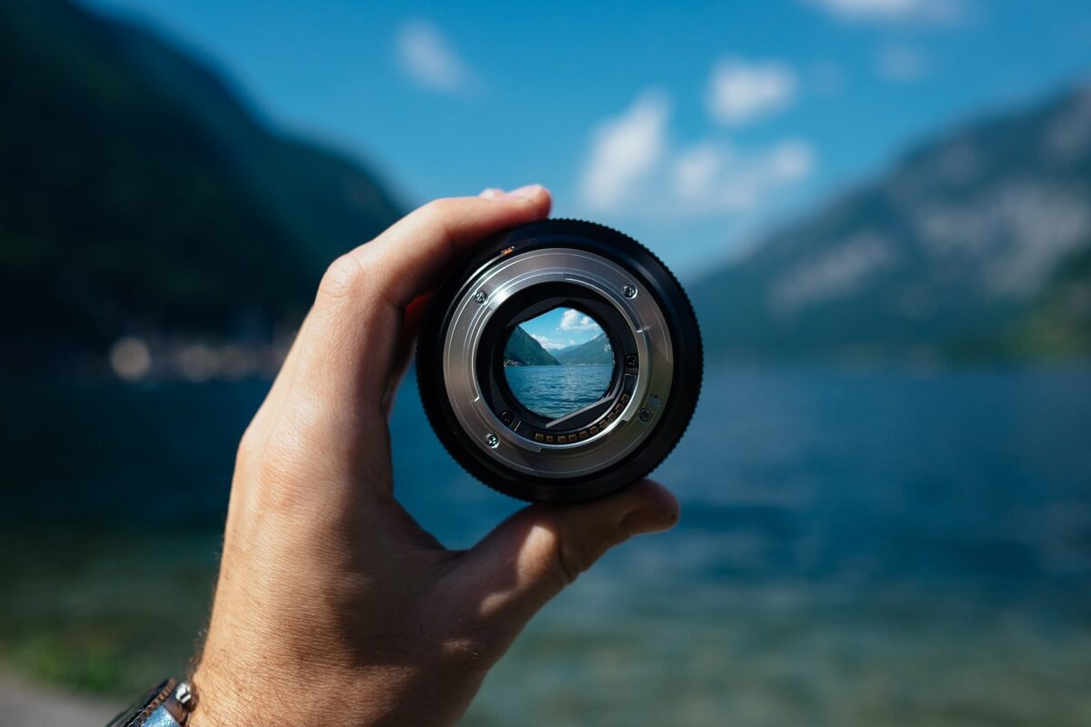 Mano agarrando lente fotográfico contra paisaje de un lago y montañas