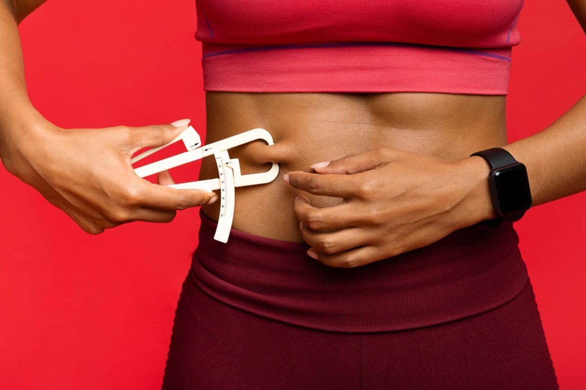 Close up of afro woman in sportswear measuring her body fat with fat caliper, red background