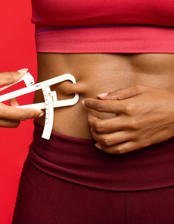 Close up of afro woman in sportswear measuring her body fat with fat caliper, red background