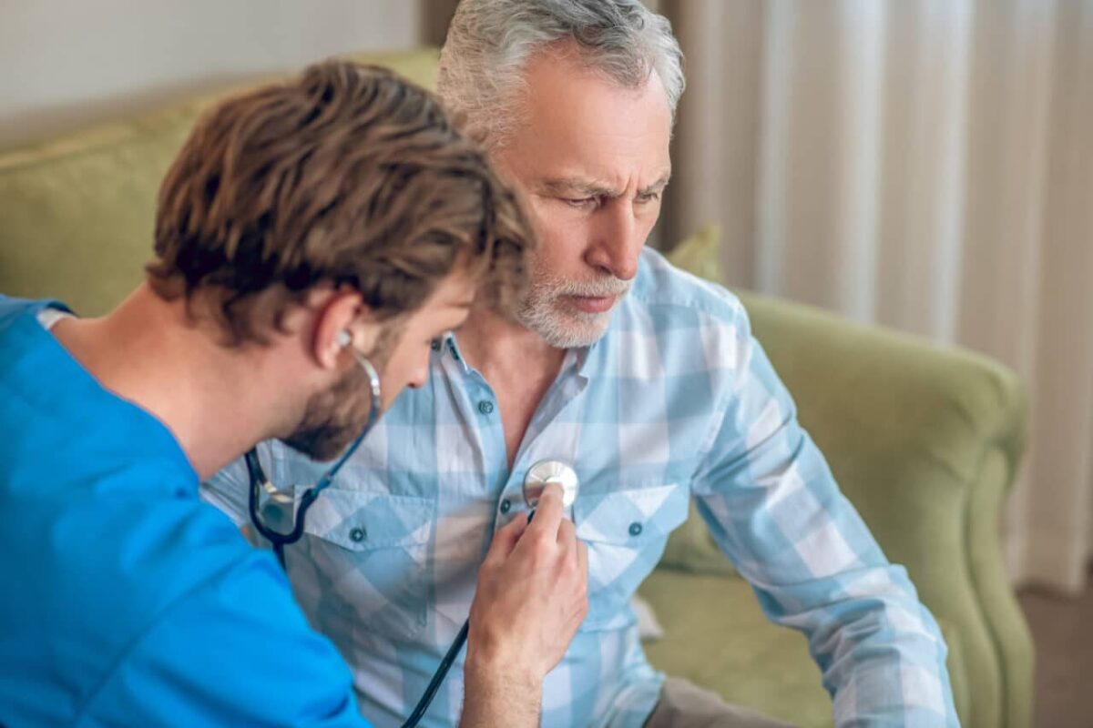 Dark-haired bearded Caucasian healthcare professional listening to the senior male patient heartbeat using a stethoscope