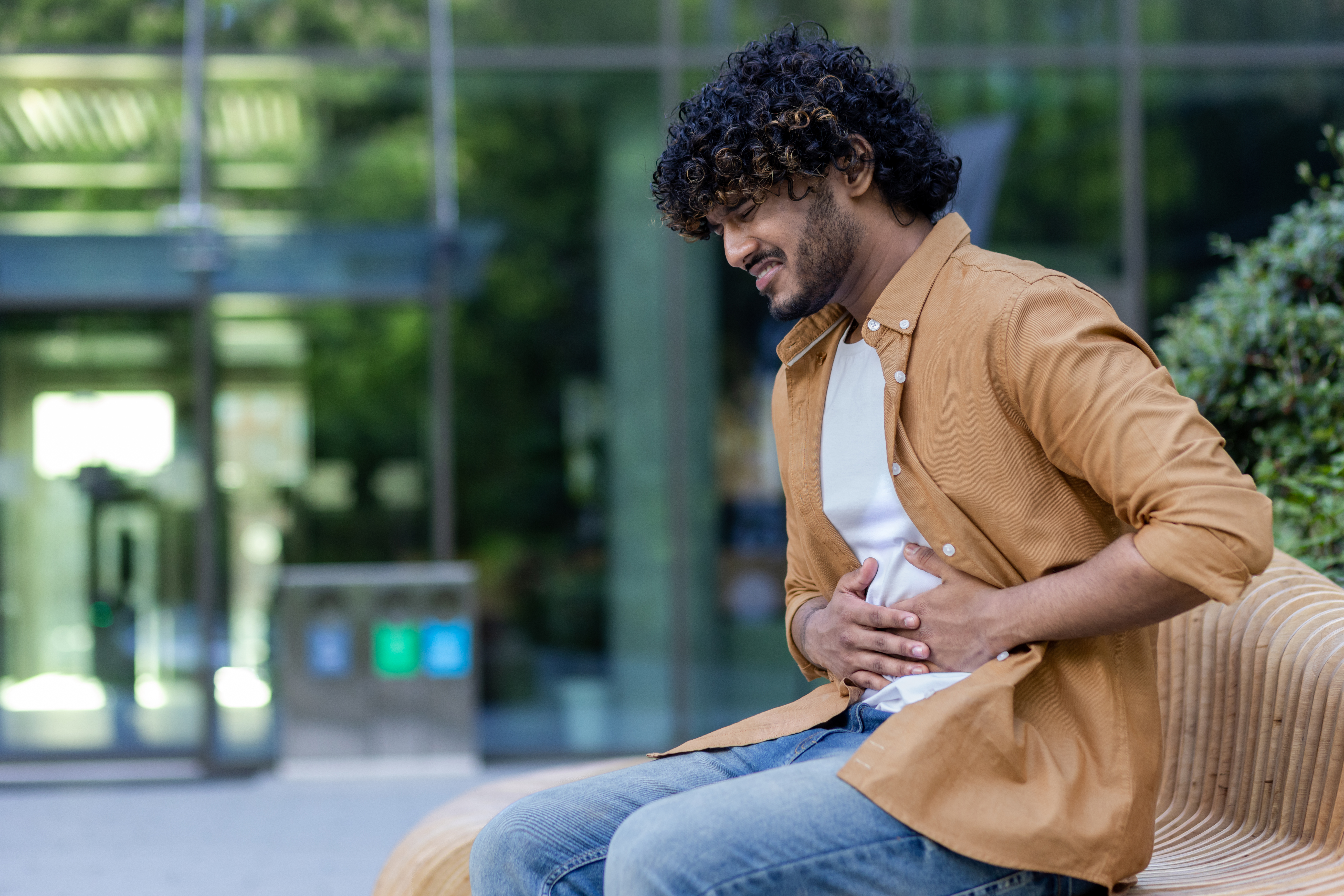 A young Indian man sits outside on a bench and suffers from stomach discomfort, holding the side of his body, grimacing in pain.